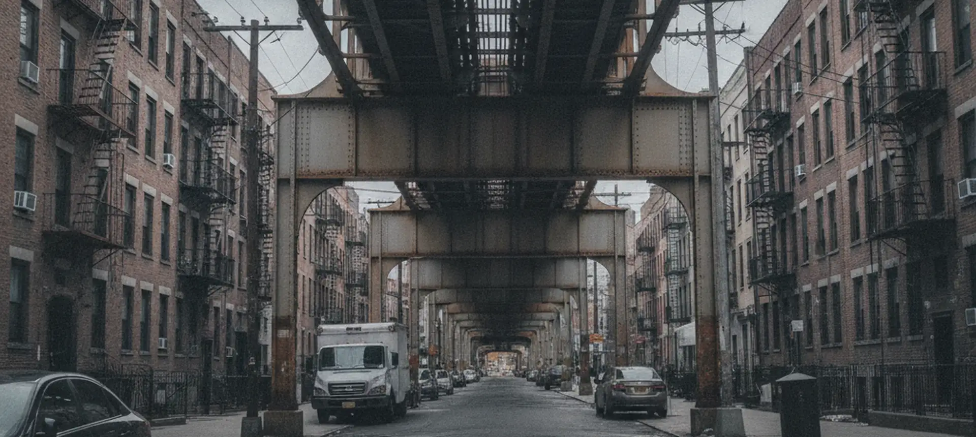 A wide view along a Bronx avenue under an elevated subway structure in deep perspective, brick apartment buildings on both sides.